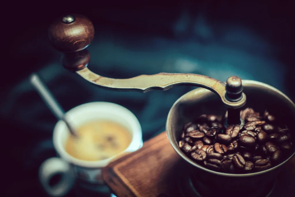 Manual coffee grinder filled with top espresso beans next to a fresh cup of espresso with golden crema.