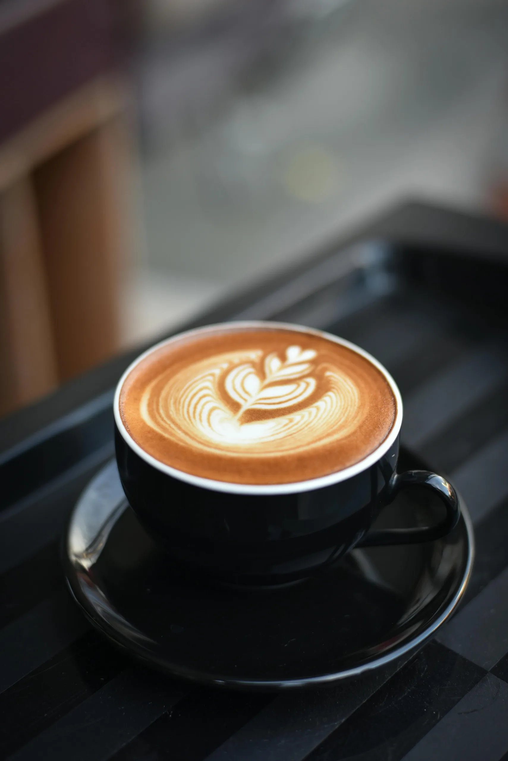 A cup of cappuccino with detailed latte art served in a black cup and saucer on a dark table.